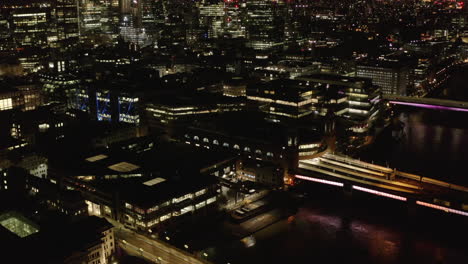 Fly-above-Thames-river-at-night.-Aerial-view-of-buildings-on-waterfront-around-Cannon-Street-train-station.-Tilt-up-reveal-of-downtown-skyscrapers.-London,-UK