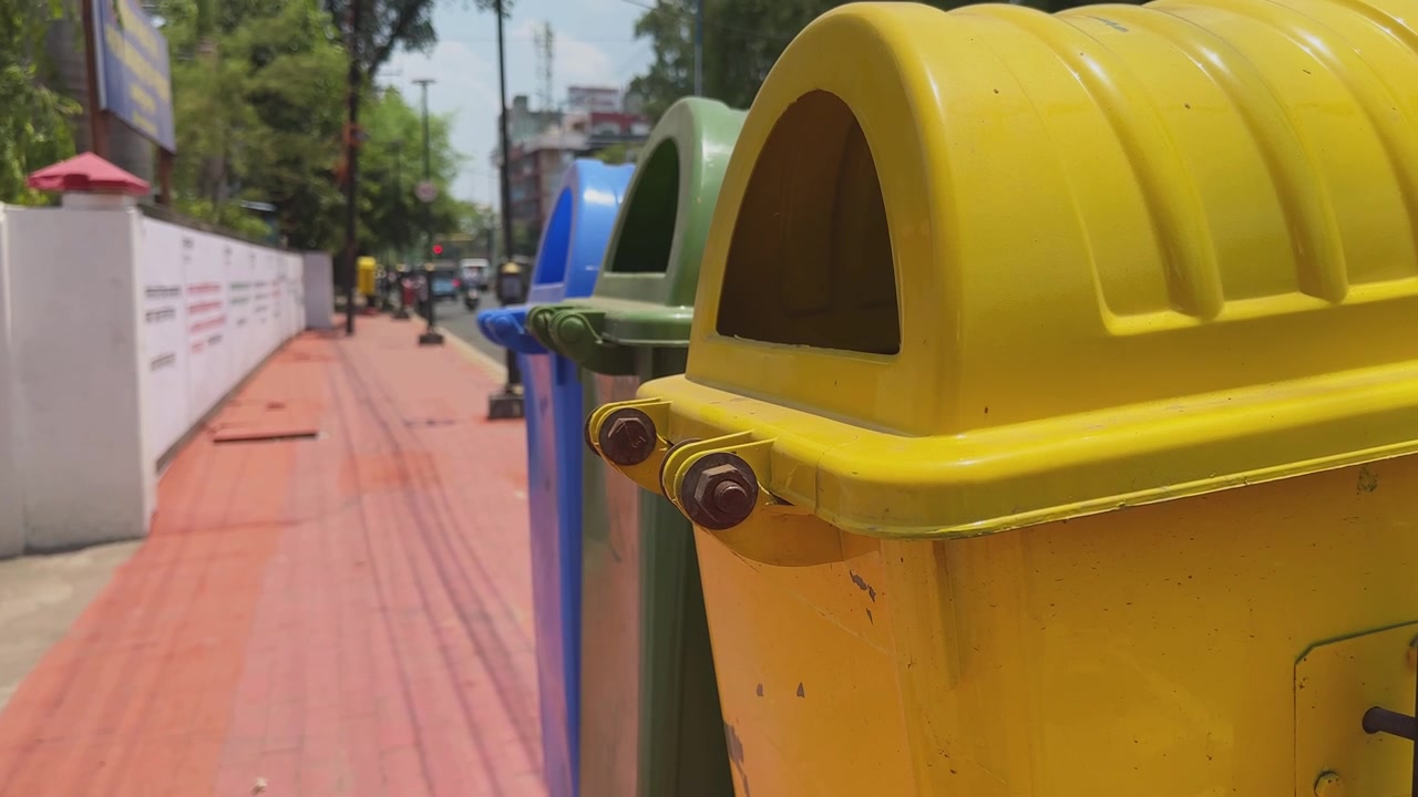 Cámara se mueve hacia una fila de coloridos contenedores de basura públicos a lo largo de una acera de la ciudad, destacando la segregación de residuos, la limpieza urbana y la infraestructura de reciclaje.