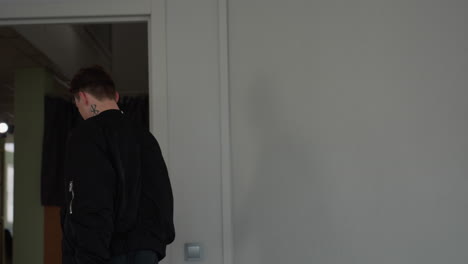 teenager wearing black jacket and belt walks forward inside bright room pulling wheeled travel bag by handle to front, soft daylight through large windows illuminates neutral minimalist interior