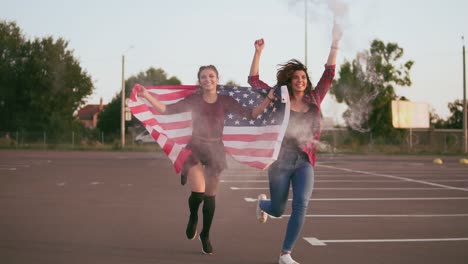 jóvenes hipster americanas felices corriendo mientras sostienen la bandera americana y sueltan granadas de humo con color blanco divirtiéndose