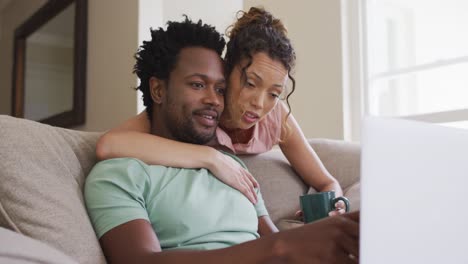 worried biracial woman with coffee embracing her male partner sitting on sofa with laptop