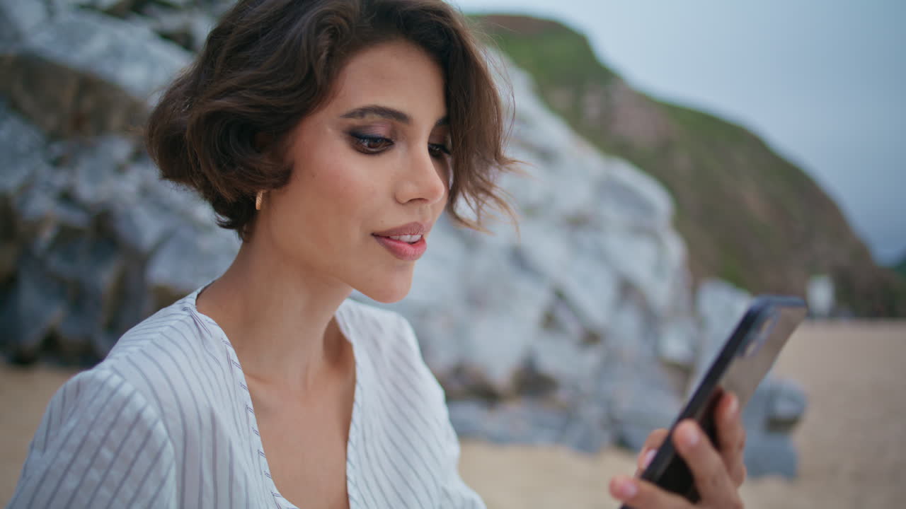 해안 근접에서 스마트폰을 사용하는 예쁜 여인. 편안한 여자가 카메라를 찾고 있습니다.