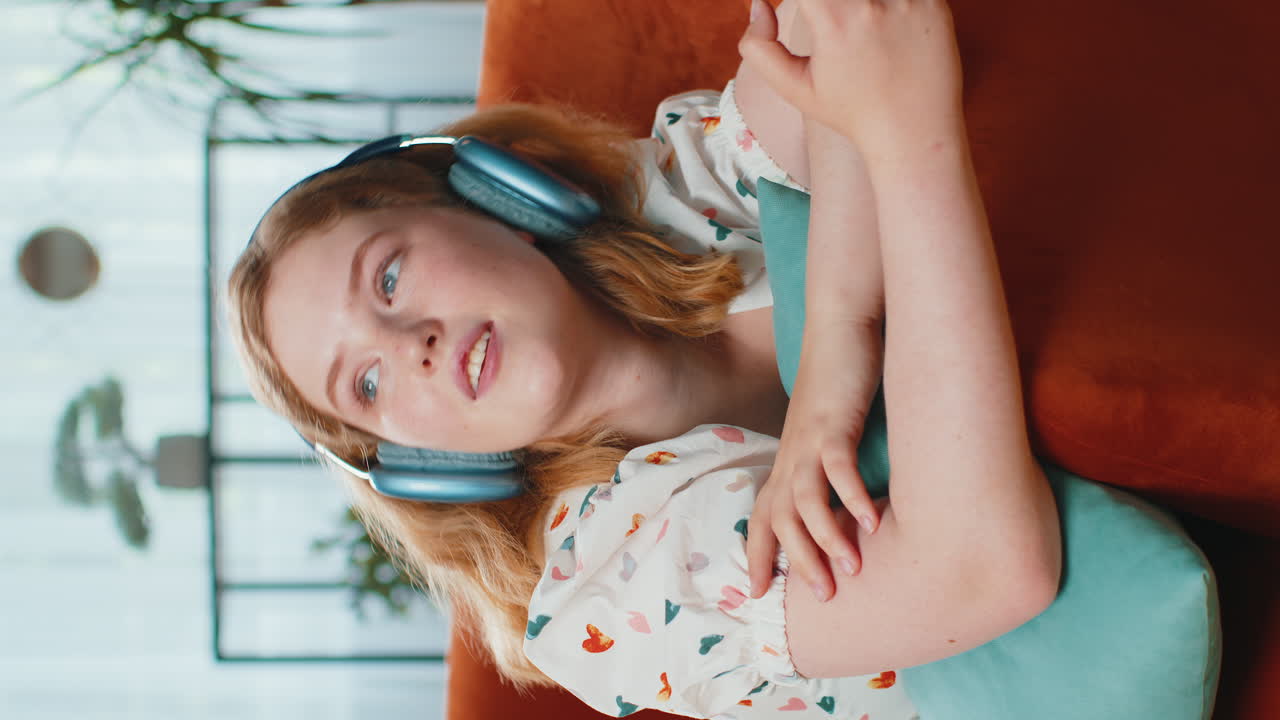 무선 헤드폰을 입은 행복한 젊은 여성이 집에서 소파에 누워서 좋아하는 음악을 듣고 휴식을 취하고 있습니다.