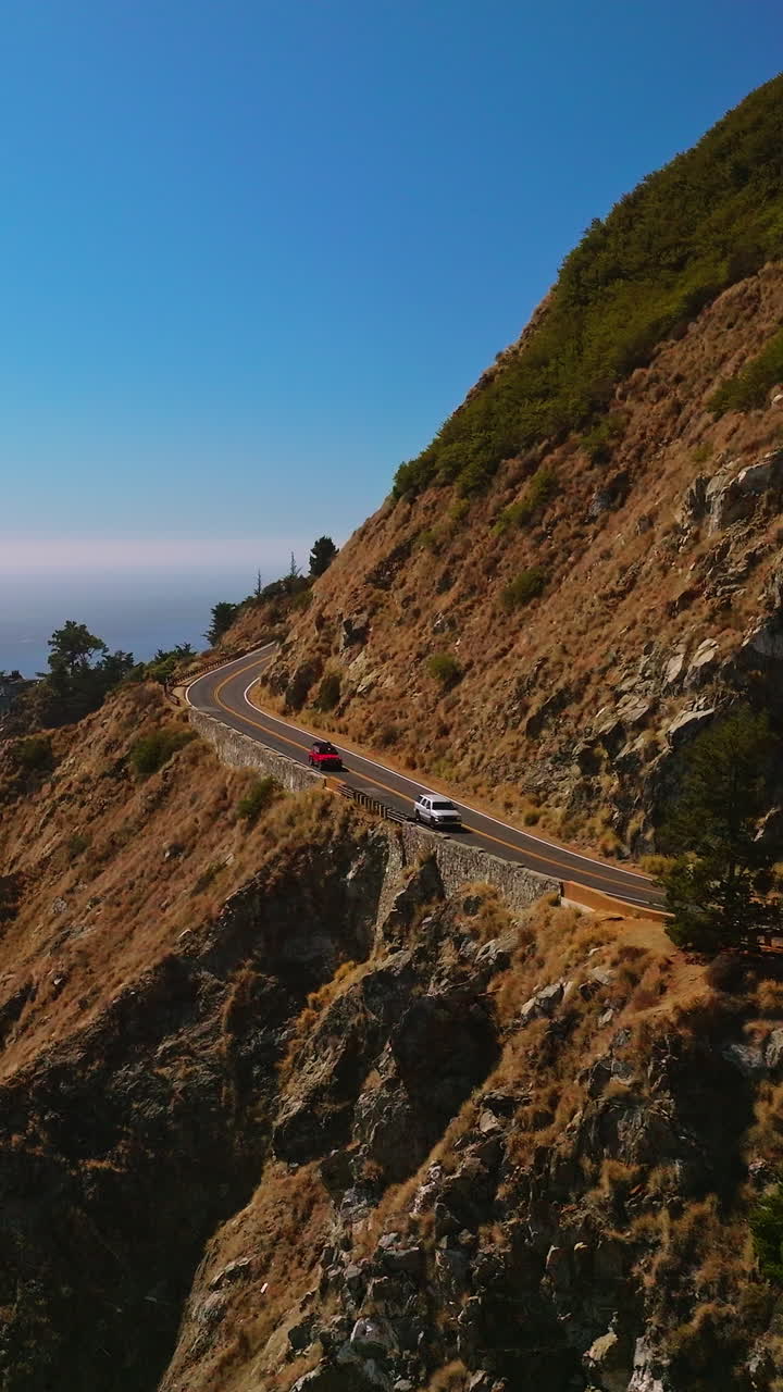 Freeway in the mountains with cars moving quickly. Beautiful rocks of Big Sur Morro Bay by the ocean at the backdrop of blue clear sky. Vertical video