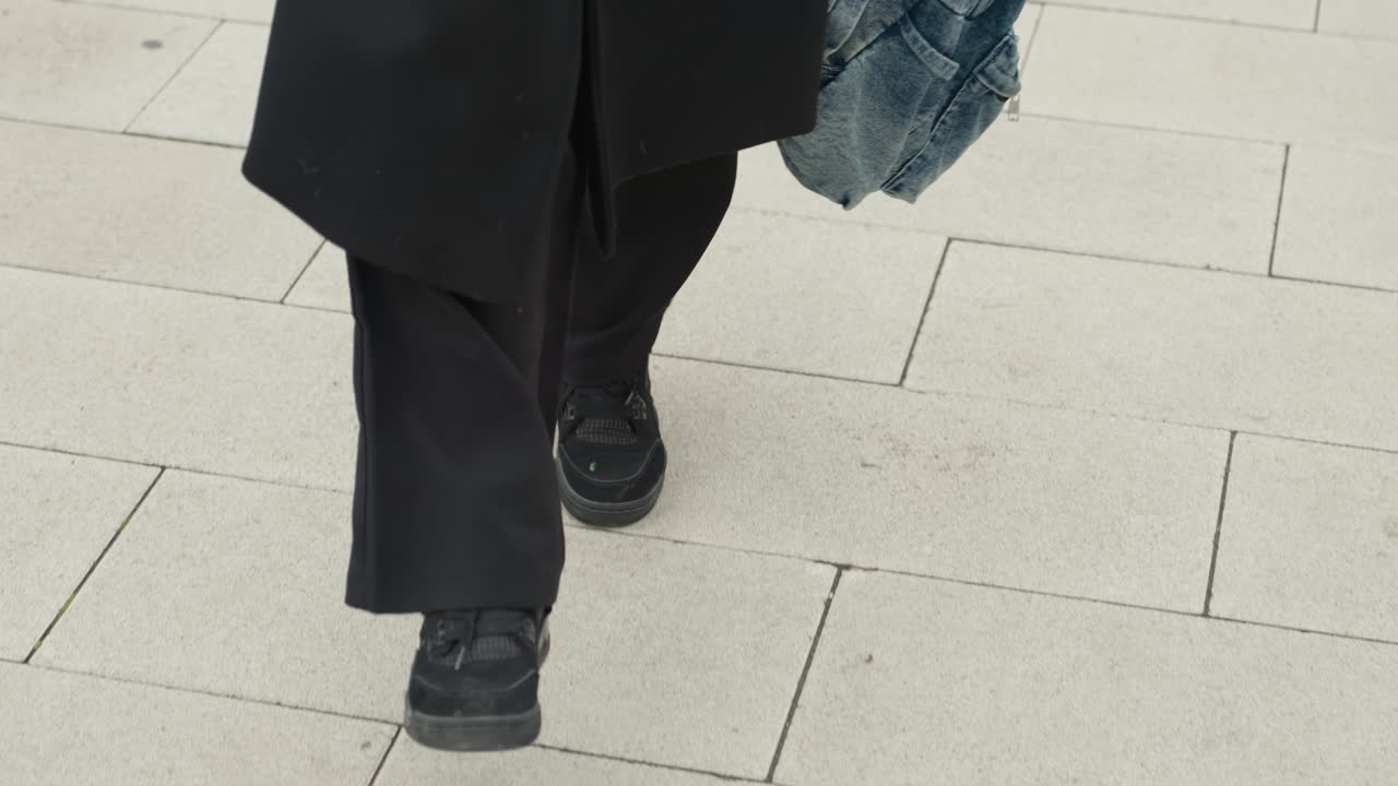 student striding with denim backpack pavement, brisk city walk lower body closeup showing black coat and trousers, swaying denim bag and decisive steps across tiled sidewalk, contemporary streetwear