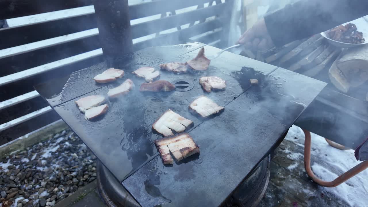Video al rallentatore di una famiglia che cucina spesse fette di pancetta di maiale all'aperto in una giornata invernale in Bucovina, nella contea di Suceava, rosolando la carne grassa direttamente sulla superficie calda della stufa in ghisa lucidata