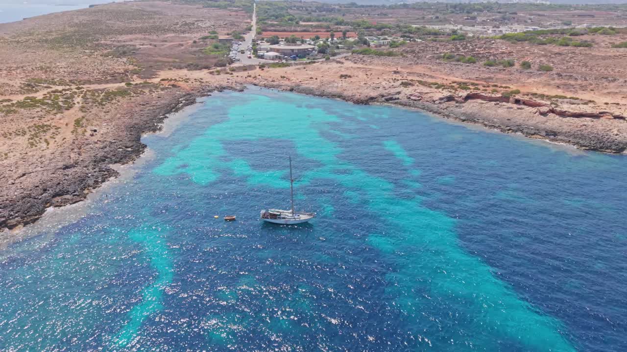 A sailboat anchored in a turquoise bay along a rocky coastline. The clear blue waters contrast beautifully with the dry, rugged landscape in the background, creating a peaceful Mediterranean scene