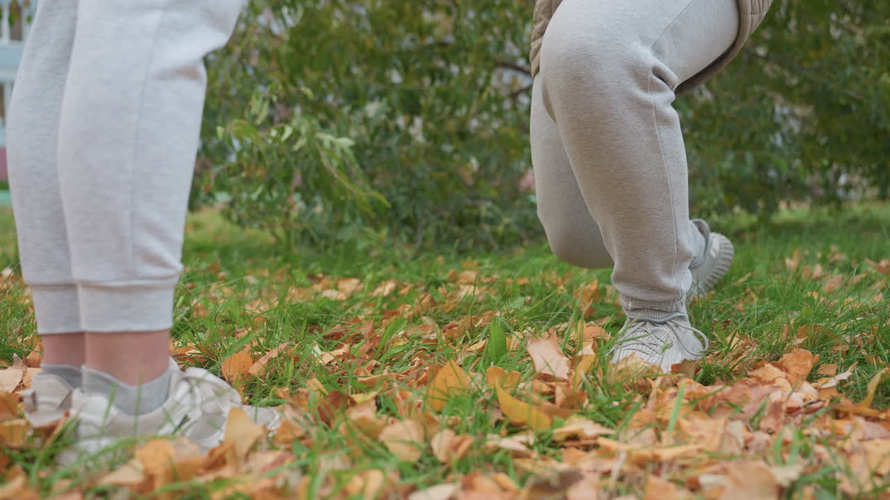 Lower body view of people in matching outfits as one gets on knees surrounded by green grass, dry autumn leaves, and distant view of building during emotional outdoor moment