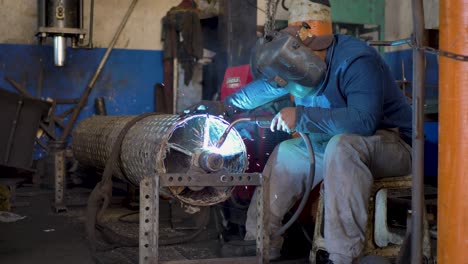 Soldador-Latinoamericano-Con-Casco-Y-Guantes-Protectores-De-Cara-Completa,-Centrado-En-Soldar-Cilindros-Metálicos-En-El-Taller.