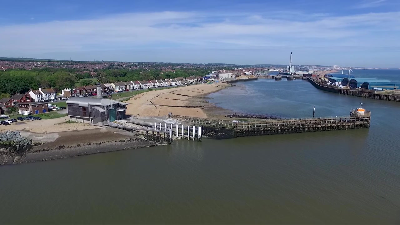 Ripresa con drone che ruota attorno a un porto rivelando una stazione per imbarcazioni, un faro, una centrale elettrica, un molo, il porto e case sullo sfondo.