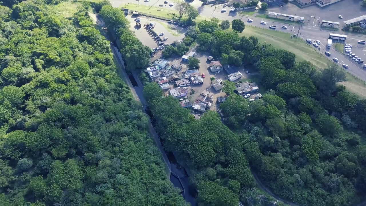drone volando lejos de un campamento de ocupantes ilegales de asentamientos informales rodeado de árboles frondosos y un canal