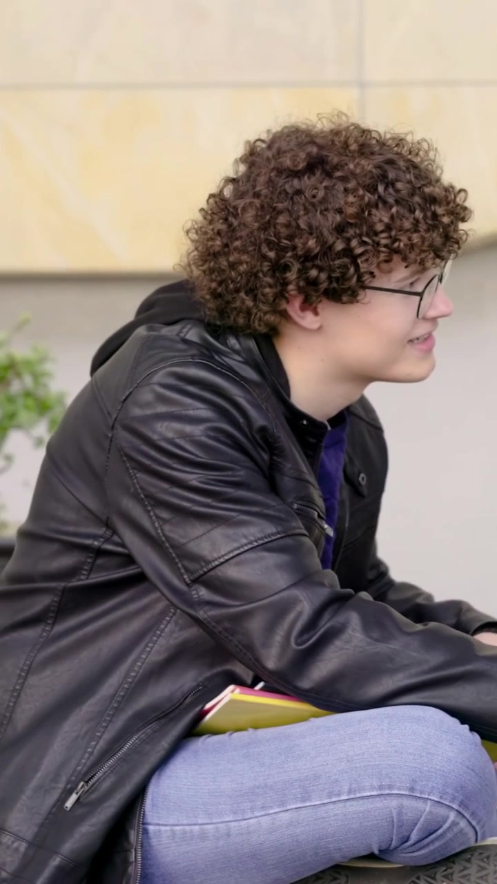 Vídeo vertical: Inclinando-se, homem de jaqueta de couro folheando cadernos em um banco do campus, estudando