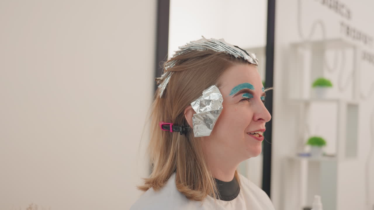 cliente di un salone con capelli avvolti in carta stagnola e ombretto blu audace, sorridente durante una sessione di colpi di sole in un moderno studio luminoso, clip per capelli che fissano la carta stagnola, atmosfera rilassata, foto profilo spontanea che mette in risalto