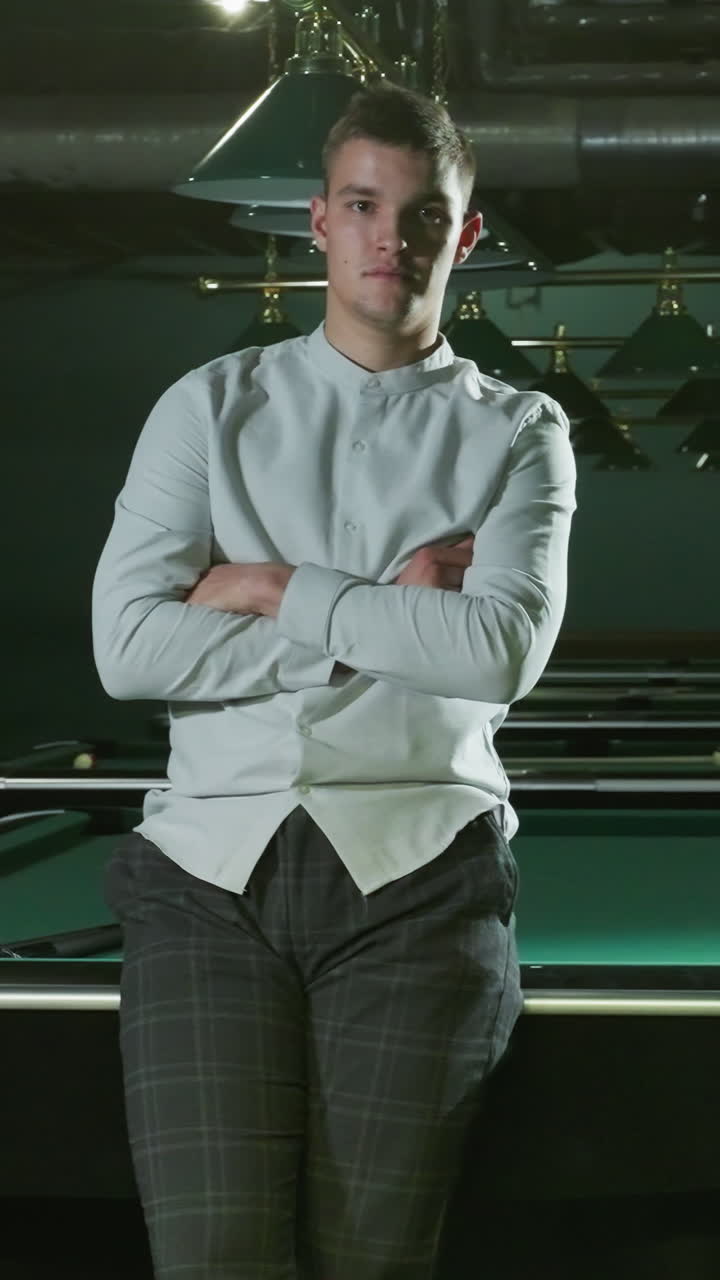 셔츠와 회색 바지를 입은 젊은이가 진지한 얼굴과 팔을 접은 녹색 빌리어드 테이블에 앉아 있습니다. 여러 개의 빌리어트 테이블, 오버헤드 램프, 그리고 현대적인 인테리어가 배경입니다.
