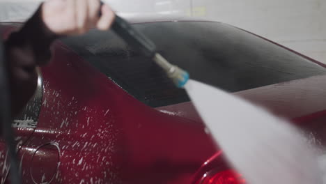 rear view of person using high pressure hose to rinse soap foam from back of red car inside professional garage environment with water streams and suds flowing dynamically over vehicle surface