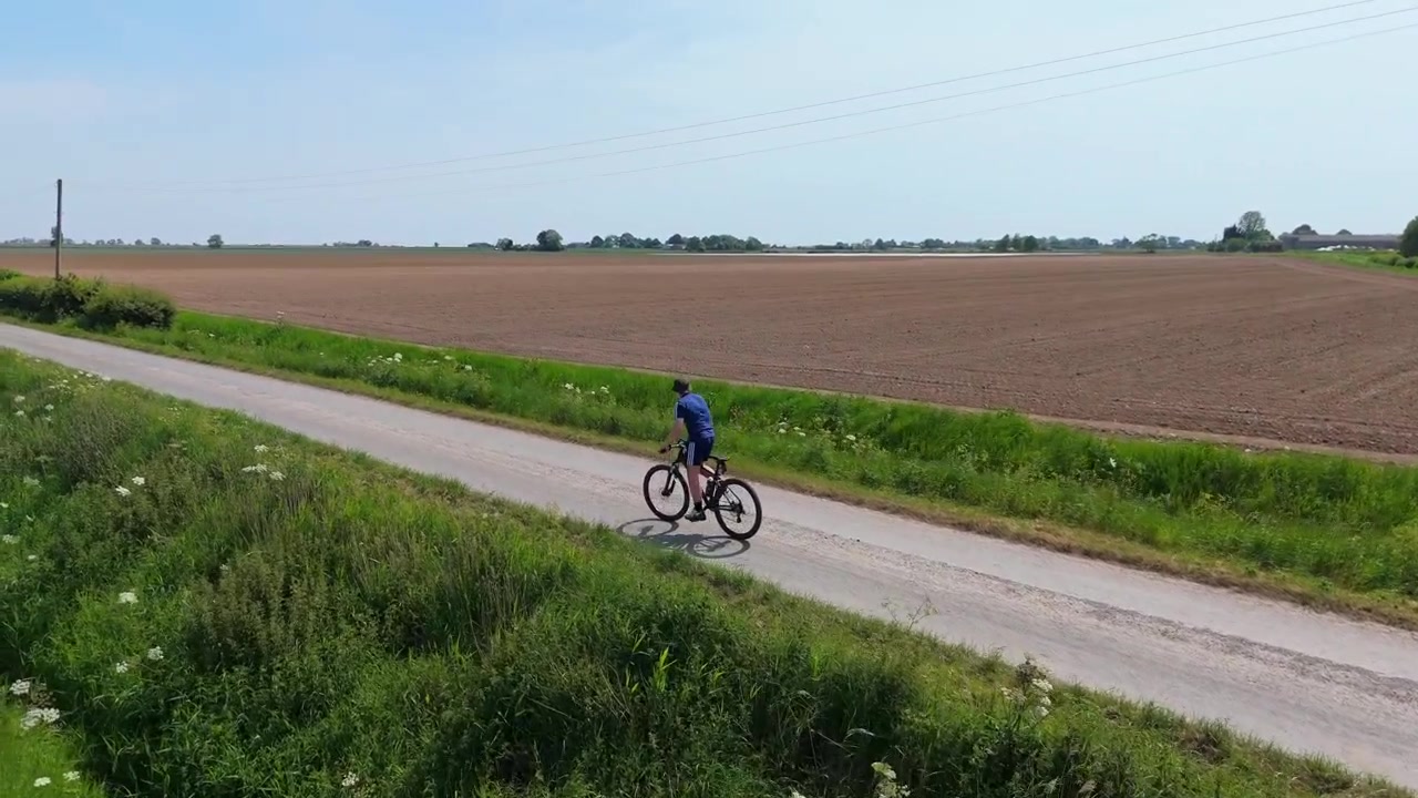 Cyklist kører på en grusvej gennem gyldne marker. Klar blå himmel omgiver det fredfyldte landskab. Frodigt grønt græs omkranser stien, med blomstrende vilde blomster.