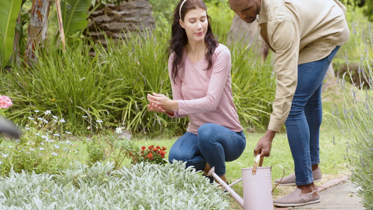 Casal multirracial jardinando juntos, mulher ajoelhada e homem segurando um regador.