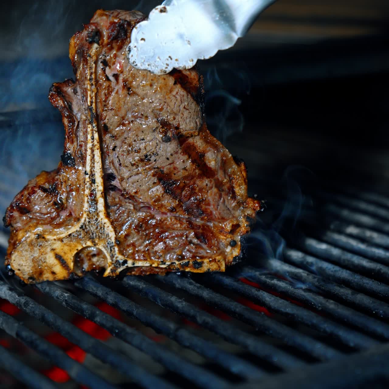 Meat on the bone is cooked on the grill. Gloved hand turns the piece of beef with tongs. Vertical screen. Close up