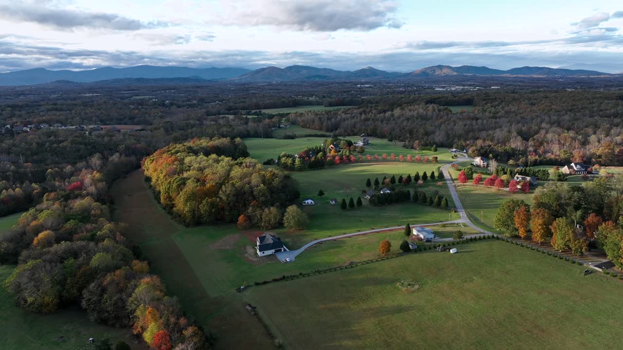 Aerial view of a peaceful American rural landscape with autumn foliage, open fields, scattered homes and distant mountains under soft evening light. Long driveway or property In USA