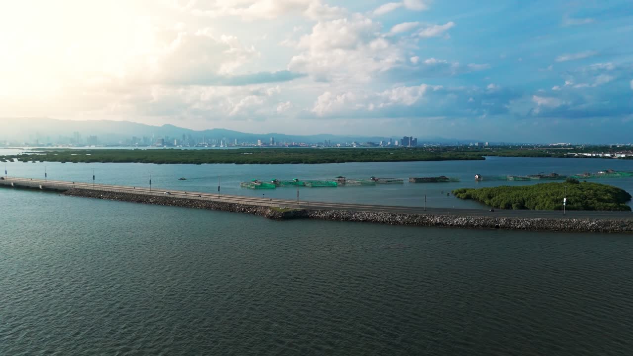 Wide drone shot of a scenic coastal road with fish farms, lush mangroves, and an urban skyline under a dramatic sky in the Philippines