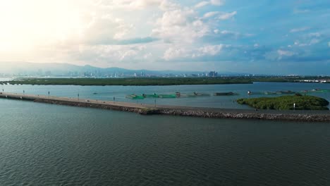 wide drone shot of a scenic coastal road with fish farms, lush mangroves, and an urban skyline under a dramatic sky in the philippines