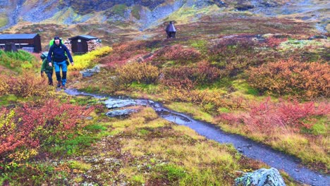 Two-hikers-with-backpacks-leaving-a-mountain-cabin-in-Abisko-national-park-in-Sweden,-walking-past-the-camera-in-a-rainy-but-colourful-autumn-terrain