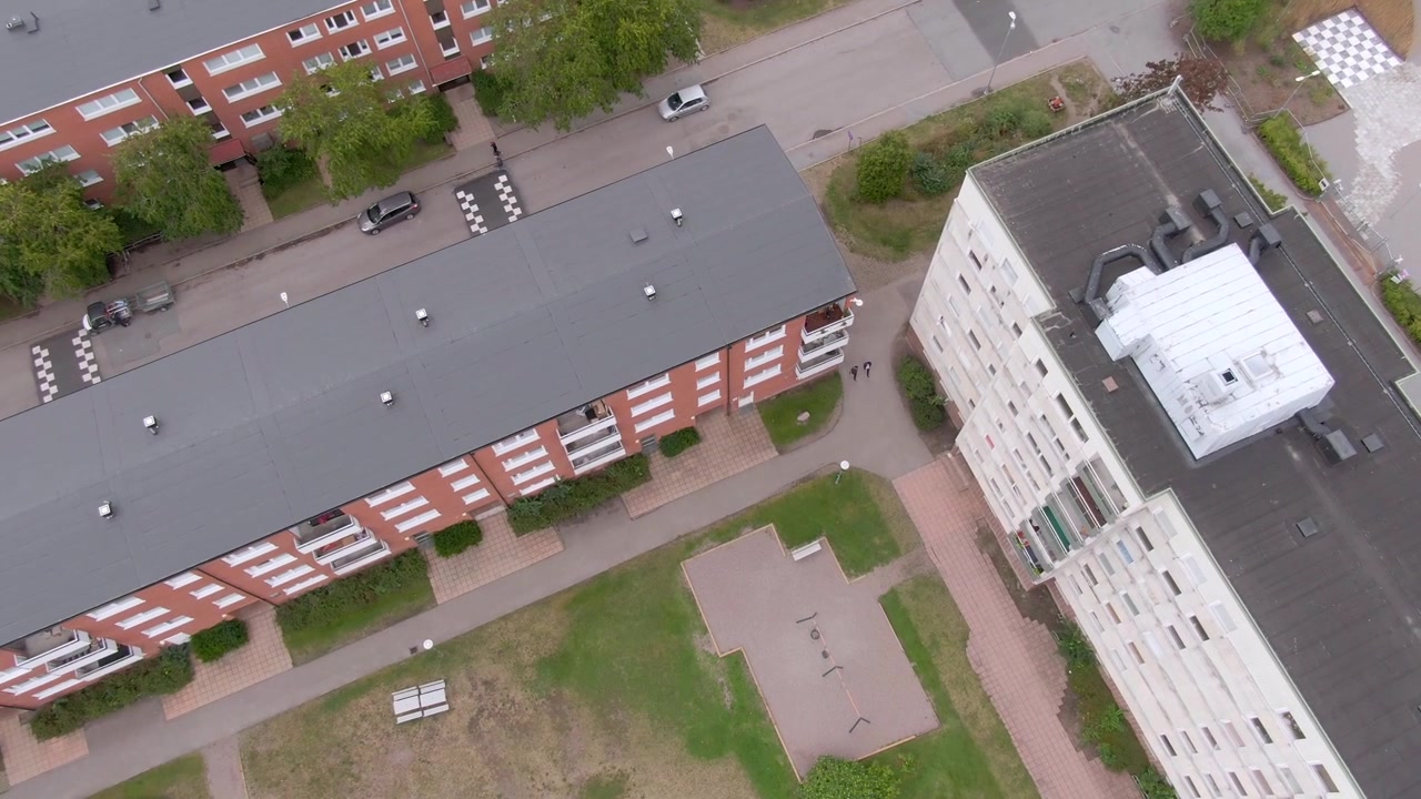 Vista zenital de telhados de apartamentos de tijolos vermelhos, unidade de HVAC, pátio gramado com caixa de areia, bancos e carros na rua