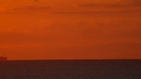 a cargo ship travels across in the distance across the ocean horizon under a glowing orange sky