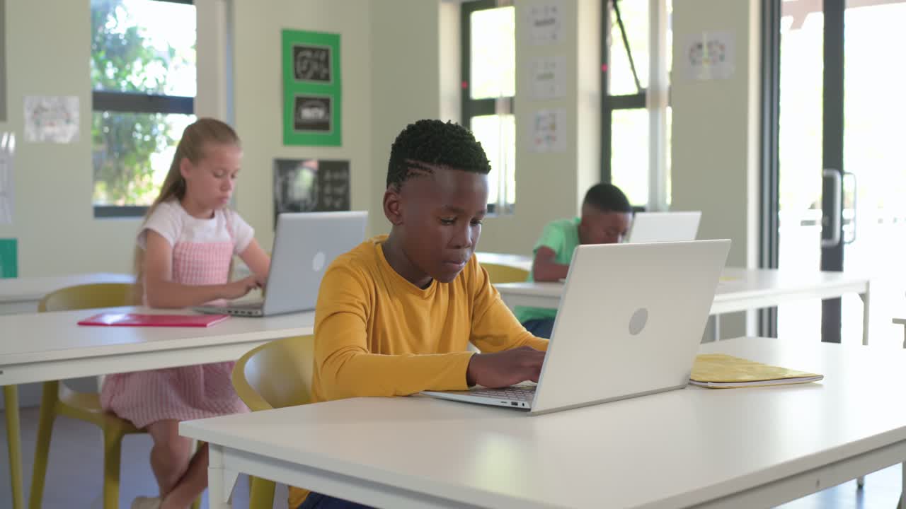 Diverse classroom classmates watching boy in yellow typing on laptop tiring leaning resting on desk