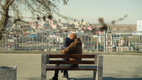 cerca de un padre sosteniendo a su hijo afectuosamente en su pierna mientras se sientan juntos en un banco, ambos con chaquetas y gorras, con vistas a un paisaje urbano con un suave y reconfortante abrazo