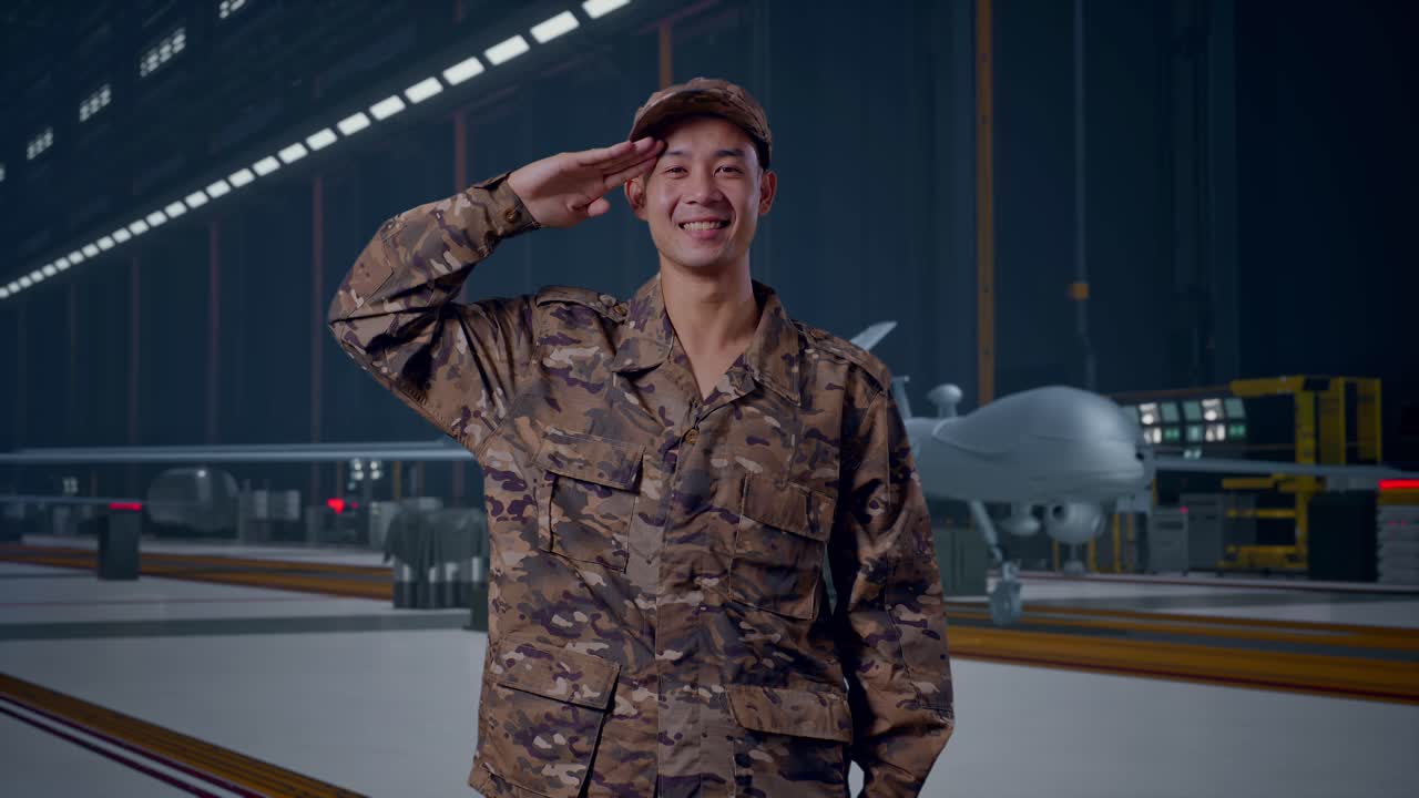 Soldato uomo asiatico che saluta militarmente e sorride mentre è in piedi accanto a un drone militare moderno in un hangar di manutenzione.