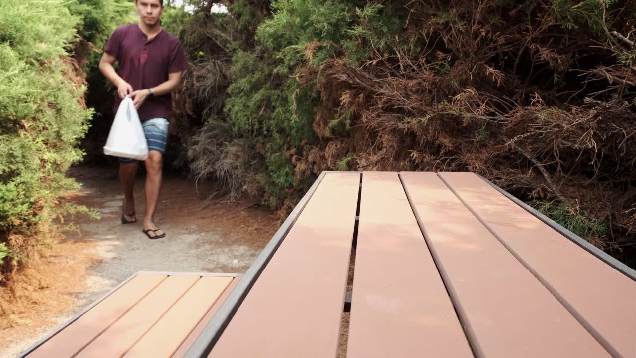 giovane maschio caucasico in abbigliamento casual che si avvicina al tavolo del parco per mettere cibo da asporto in un sacchetto di plastica