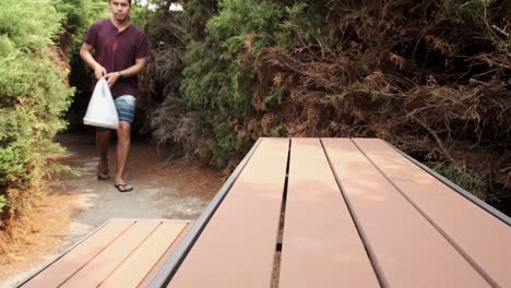 joven masculino caucásico en ropa casual que se acerca a la mesa del parque para colocar comida para llevar en una bolsa de plástico
