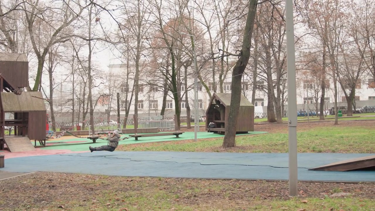 Parco giochi vuoto con struttura in legno in una mattina d'autunno, foglie marroni sparse sulla superficie di sicurezza blu, alberi spogli ed edifici residenziali distanti, atmosfera tranquilla del parco, attrezzature comunali usurate.