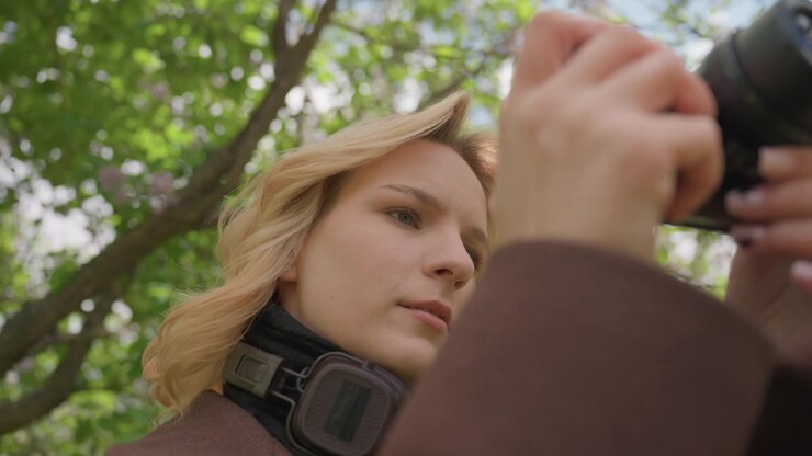 Close up shows lady outdoors using binoculars to observe nature. Woman adjusts device focusing on details like her blonde hair and headphones while surrounded by greenery