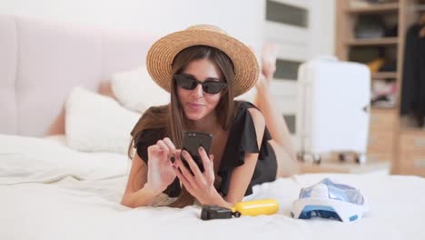 hermosa chica en traje de baño y sombrero acostado en la cama elige por teléfono dónde volar de vacaciones, en el fondo de una maleta recogida