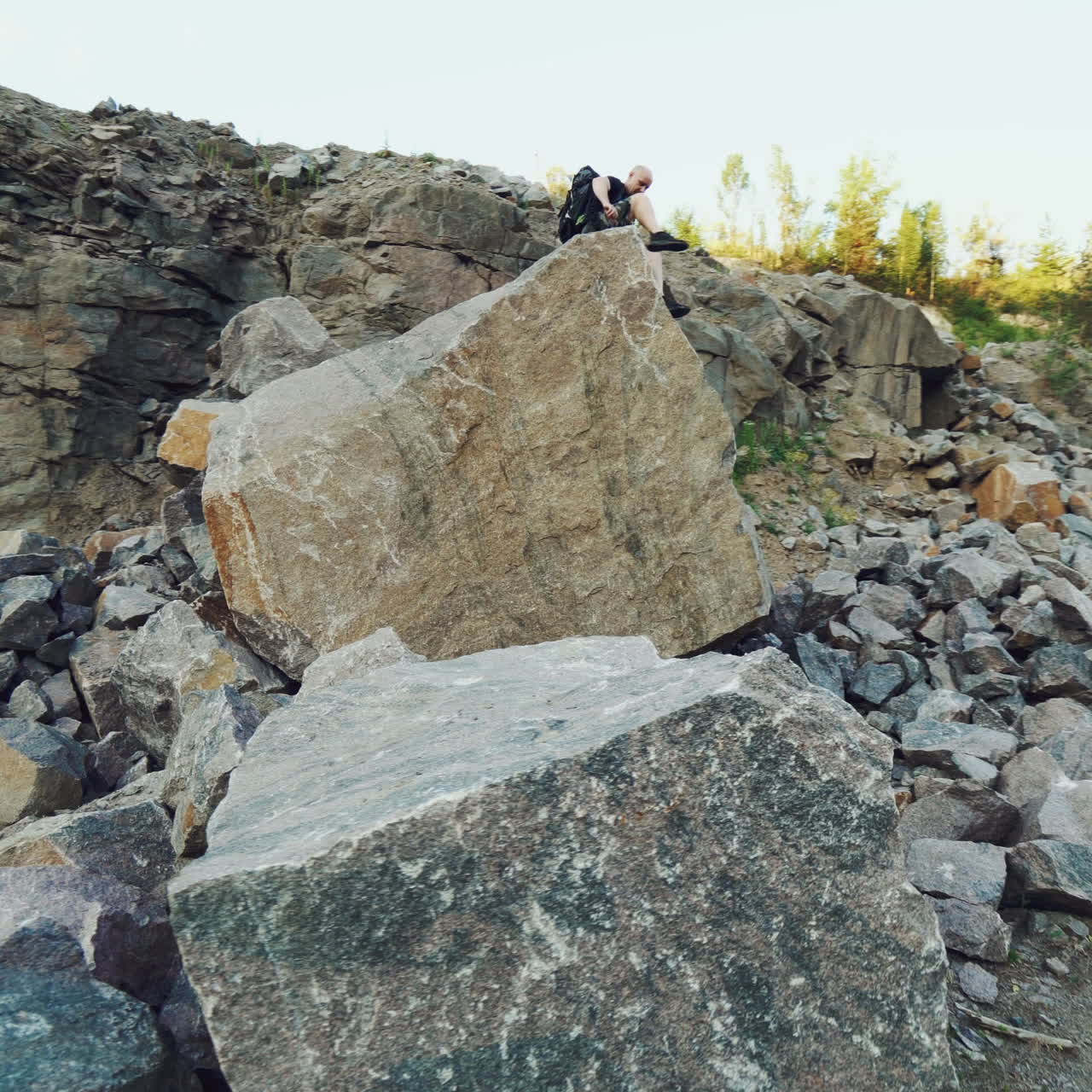 A traveler in shorts and with a backpack climbs onto a huge stone and sits on it amid a quarry with a lake in the center.