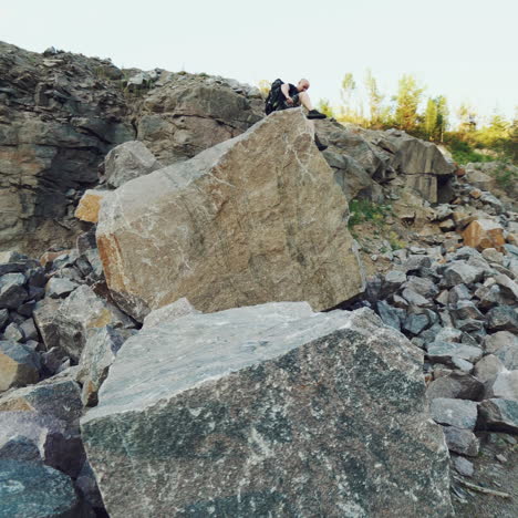 a traveler in shorts and with a backpack climbs onto a huge stone and sits on it amid a quarry with a lake in the center.