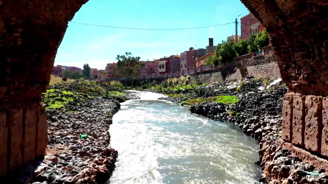 Toma HD del río Oum Er-Rbia fluyendo por la ciudad de Jenifra, visto a través de un arco de piedra. Presenta agua corriente, orillas rocosas y arquitectura urbana en el Atlas Medio.