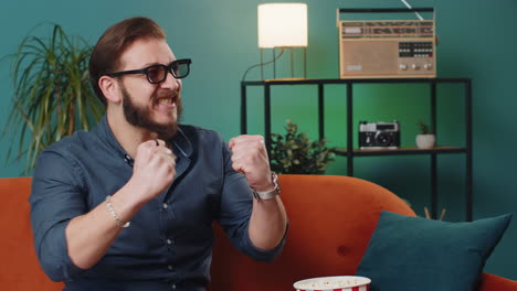 lebanese man sits on sofa eating popcorn watching interesting tv serial sport game online at home