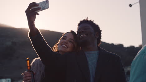 Eine-Gruppe-Von-Freunden-Hängt-Zusammen-Ab-Und-Genießt-Die-Party-Auf-Dem-Dach-Bei-Sonnenuntergang,-Macht-Selfie-Fotos-Mit-Dem-Smartphone,-Trinkt-Alkohol-Und-Hat-Spaß-Am-Wochenende-Beim-Gemeinsamen-Feiern