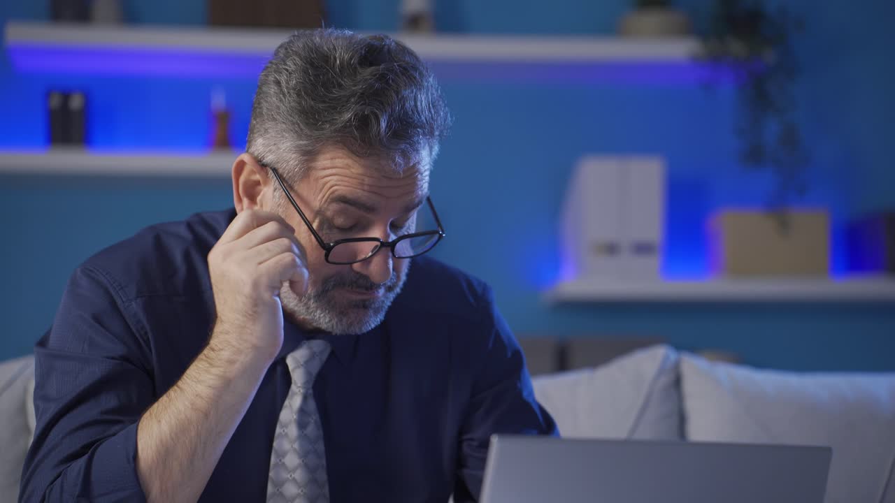 Mature businessman works alone at night at home, he gets very tired and sleepy.