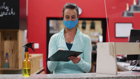 Mujer-Caucásica-Con-Mascarilla-Detrás-Del-Mostrador-En-La-Recepción-Del-Gimnasio-Pasando-El-Portapapeles