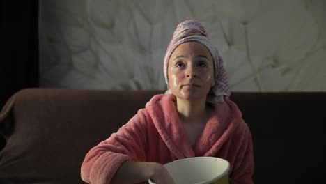 woman watching a late night movie at tv, eating popcorn. bathrobe, facial mask