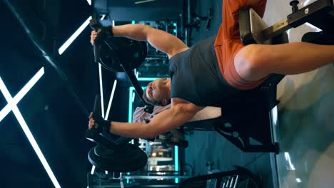 A muscular Caucasian man performs an incline bench press in a modern gym, wearing orange shorts and a sleeveless shirt, displaying a strong and toned upper body - vertical