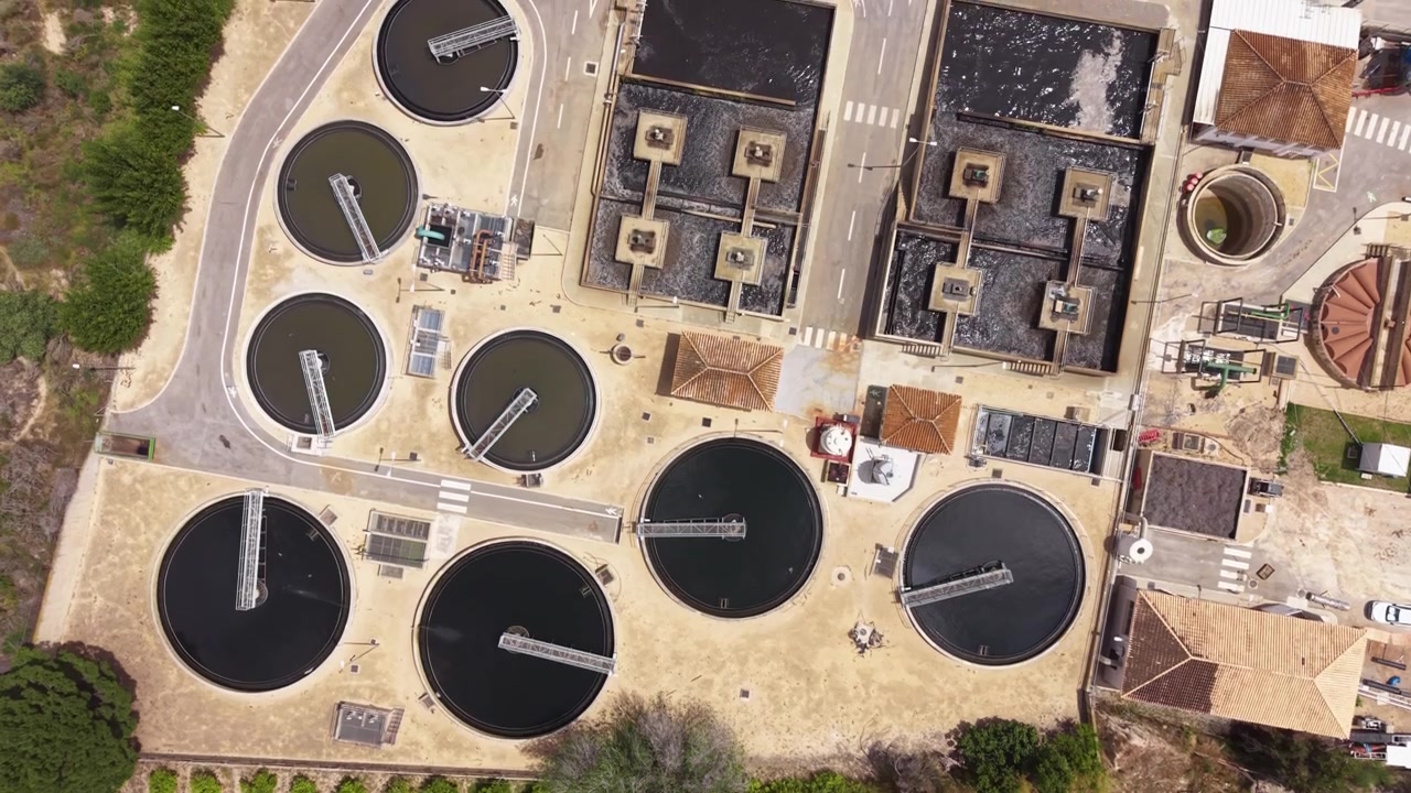Overhead view of an urban wastewater treatment plant featuring circular settling tanks, rectangular clarifiers, and industrial buildings surrounded by vegetation