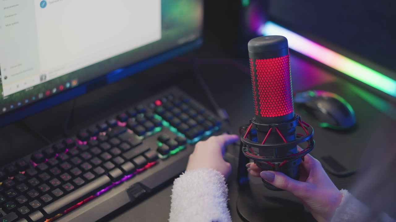 lady in fluffy jacket brings red lit studio microphone forward and adjusts cable beside glowing rgb light strip keyboard monitor setup ready for streaming gaming recording in cozy tech workspace