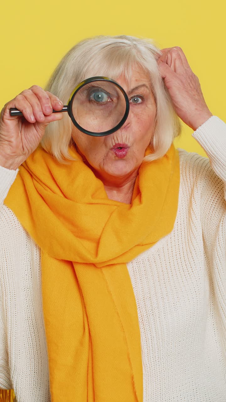 Vecchia donna anziana tiene una lente d'ingrandimento vicino al viso guardando la telecamera con un grande occhio zoomato che analizza