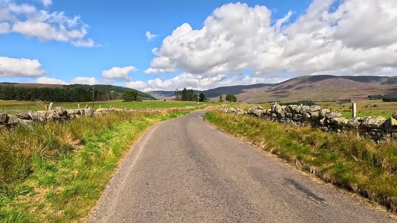Point-of-view drive down a narrow country road bordered by stone walls, with rolling hills, green fields, and bright daylight under scattered clouds