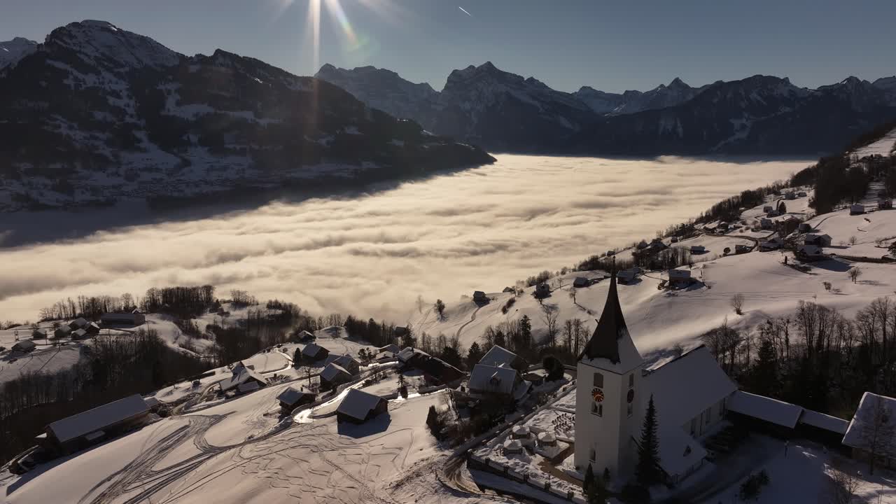 Aerial view of Amden, Switzerland, featuring the Church of St. Gallus on a snow-covered hillside overlooking a valley filled with dense clouds. The winter landscape, alpine houses and mountain peaks.